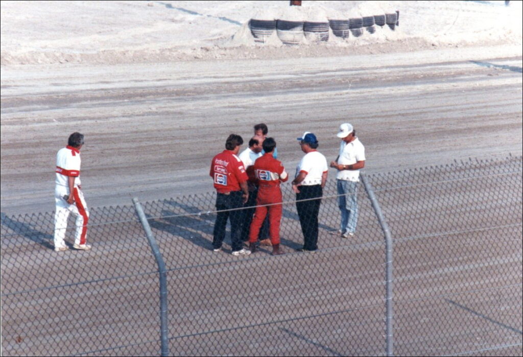 Track and event officials meet trackside to discuss the situation. The decision was made to cancel the event. Castrol Oil Advertising and Promotion Manager, Craig Hill, seen walking toward the officials. Rich Vogler in red racing suit,Harvey Hudes to his right,Myles Brandt far right, Dave Cook in the middle meet with USAC officials
